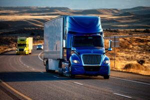 Blue tractor trailer on highway with a yellow tractor trailer and a car behind it