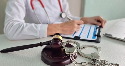 A doctor writing on a clipboard with a gavel and handcuffs placed on the desk, symbolizing medical malpractice and legal accountability.