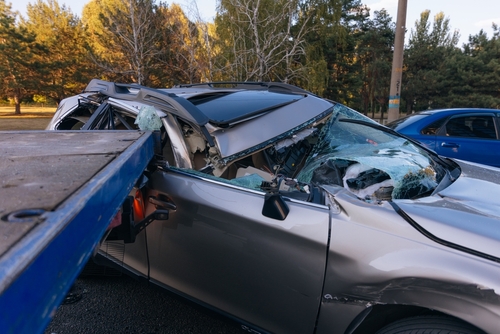 Severely damaged silver car with a crushed roof and shattered windshield being towed, likely after a serious accident, on a tree-lined road.